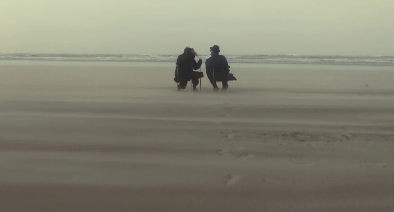 Faces Places (2017) movie still: wide — Two individuals sit on portable chairs on a desolate, windy sandy beach. The surf is visible in the distance…