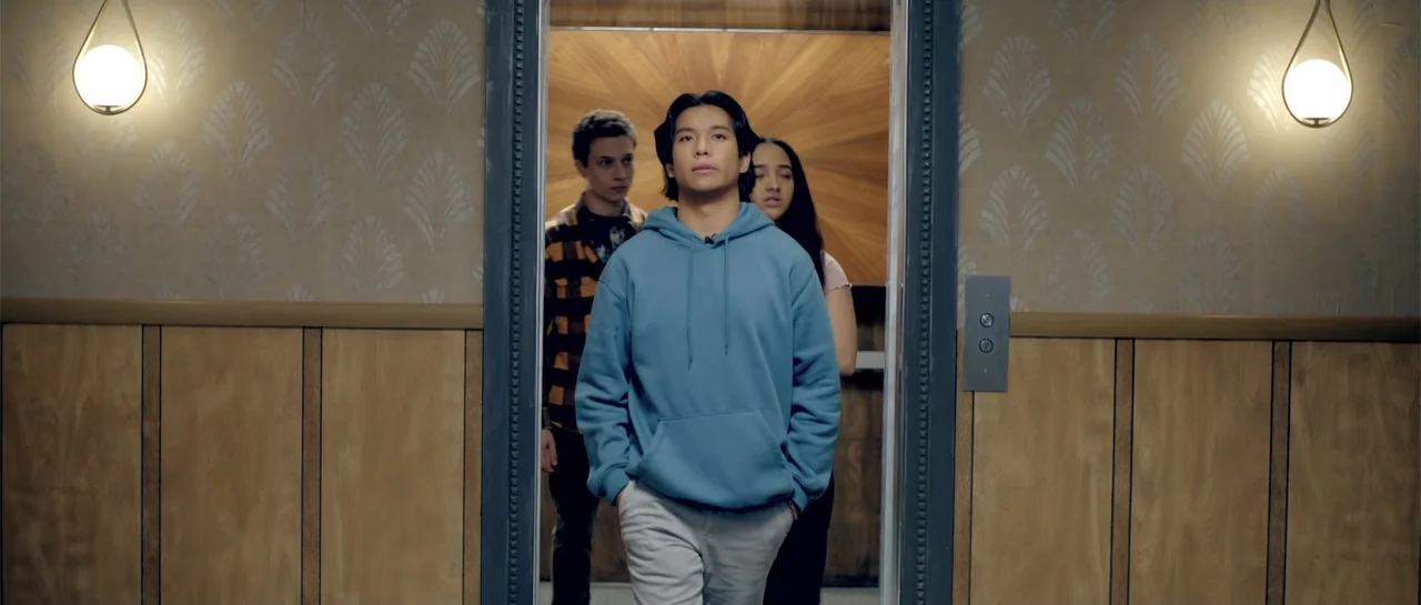 Elevator Game (2023) movie still: medium — An elevator interior features wood-paneled walls, a starburst-pattern back wall, and beige patterned…