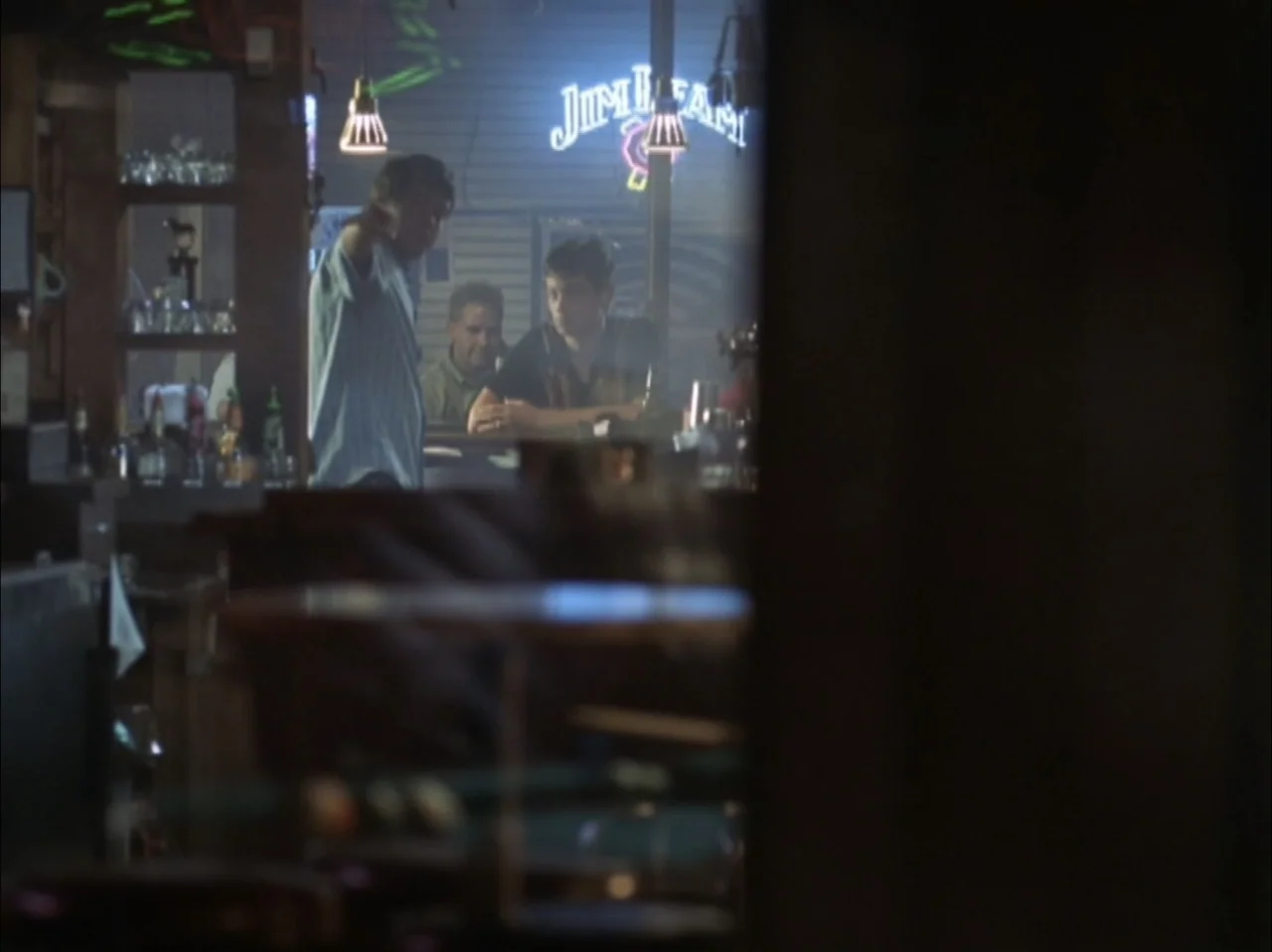 The Laramie Project (2020) movie still: medium — A dimly lit tavern counter displays a bright neon sign in the background. Two men sit at the…