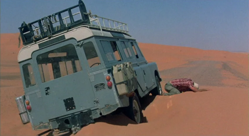 The Passenger (1975) movie still: wide — An expanse of orange sand dunes stretches toward a clear blue sky under bright sun. A light blue Land Rover…