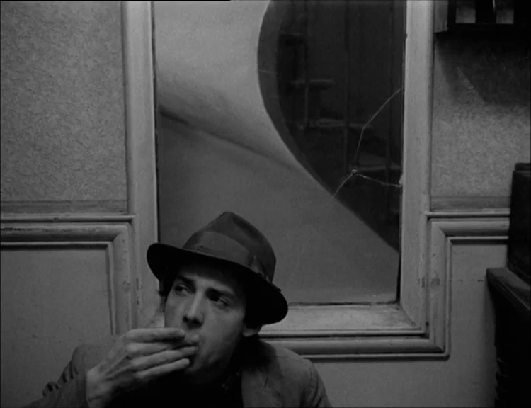 Alphaville (1965) movie still: medium — An interior space features a wall with molding and a rectangular paneled window. A man wearing a fedora sits…