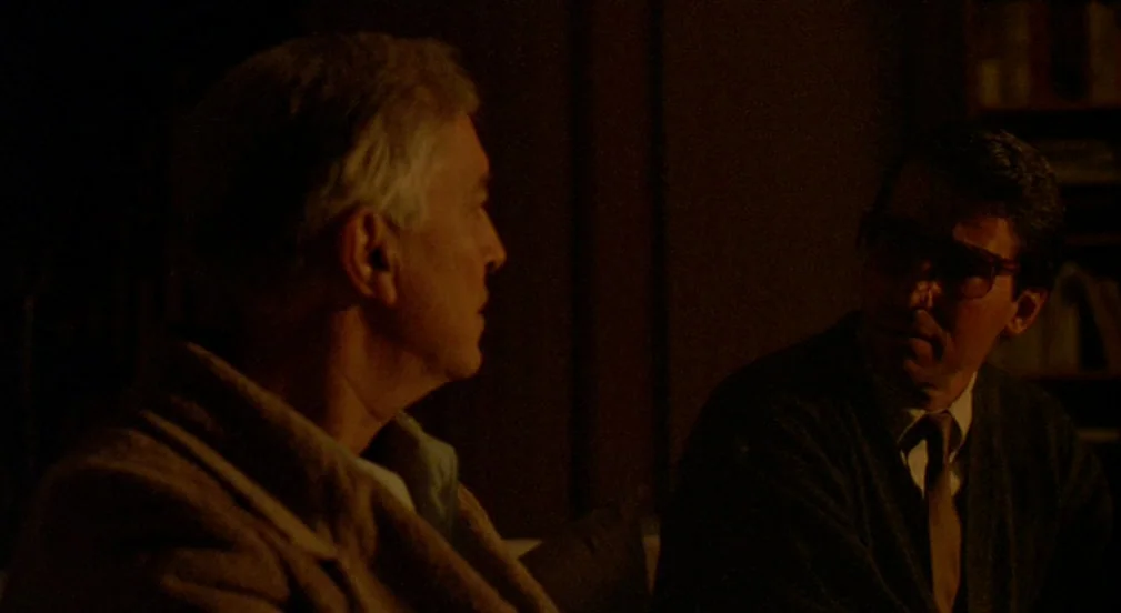 Crimes and Misdemeanors (1989) movie still: medium — A dark, dimly lit indoor space with blurred shelves visible in the background. Two men sit…