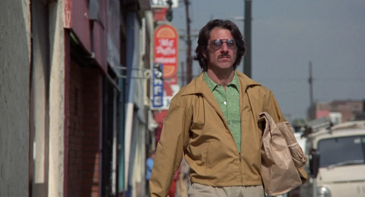 Straight Time (1978) movie still: medium — A man walks along a sidewalk lined with commercial storefronts and vertical signage. The subject carries…