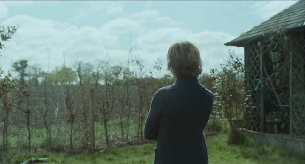 45 Years (2015) movie still: medium — A rural property features a weathered wooden outbuilding and a leafless fenced orchard area. A middle-aged…