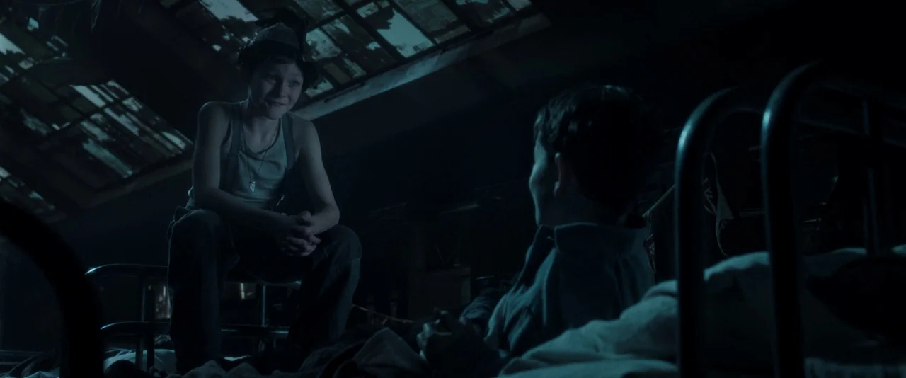 Pan (2015) movie still: medium — An attic room features a large, dark skylight with a visible metal frame structure. A boy sits on the end of a bed…
