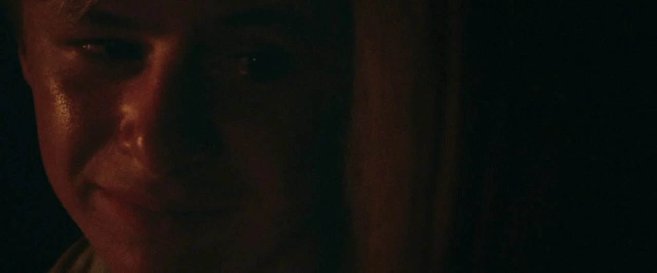 Adoration (2019) movie still: closeup — A dimly lit room holds a single person in near darkness. A young man with light hair sits while illuminated…