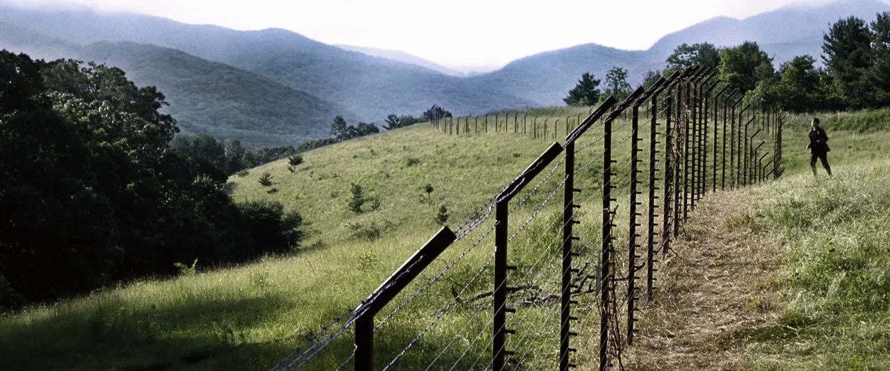 The Hunger Games (2012) movie still: wide — A vast grassy hillside is partitioned by a tall, reinforced barbed wire fence. A man in dark clothing…