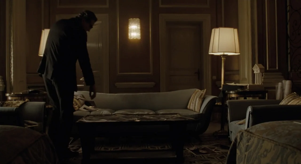 The Great Beauty (2013) movie still: medium — A dimly lit classic interior features a sofa and coffee table with scattered documents. A man in a…