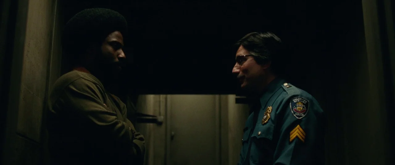 BlacKkKlansman (2018) movie still: medium — A dimly lit narrow corridor featuring dark industrial walls and shadowed corners. A man with an afro and…