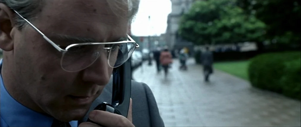 The Insider (1999) movie still: closeup — A man stands on a grey paved walkway with blurry figures in the background. The man holds a black…