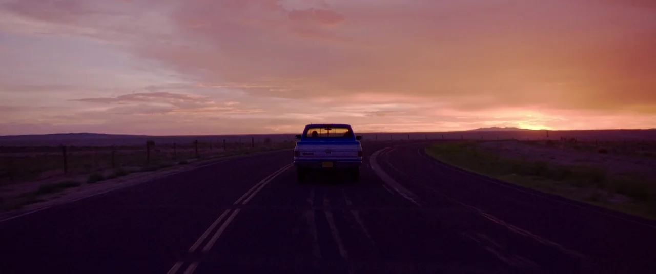 Love Lies Bleeding (2024) movie still: wide — A dark blue GMC pickup truck drives away along a two-lane asphalt road. The scene is set in an open…