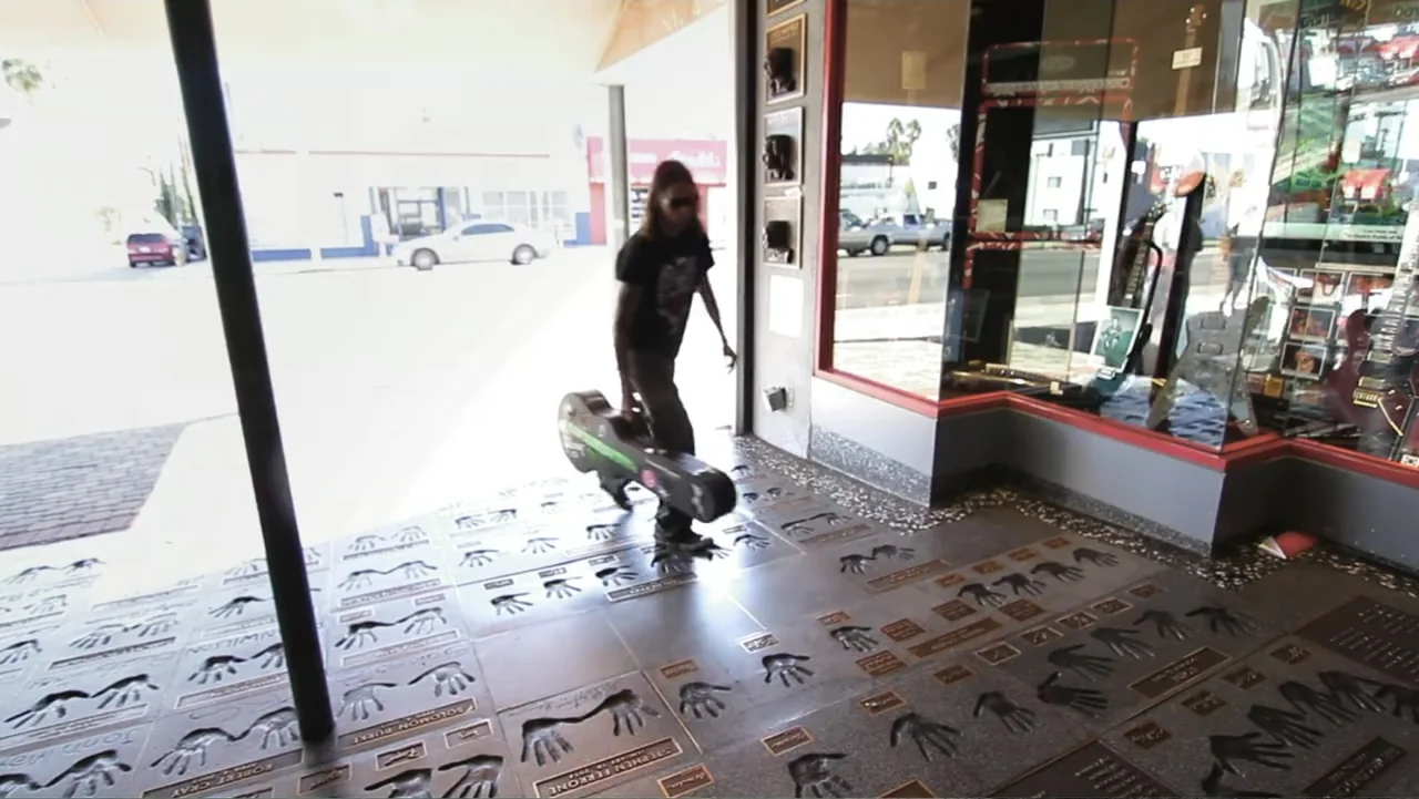 Control (2022) movie still: fullbody — A sidewalk paved with commemorative handprints leads toward an open guitar shop storefront. A man with long…