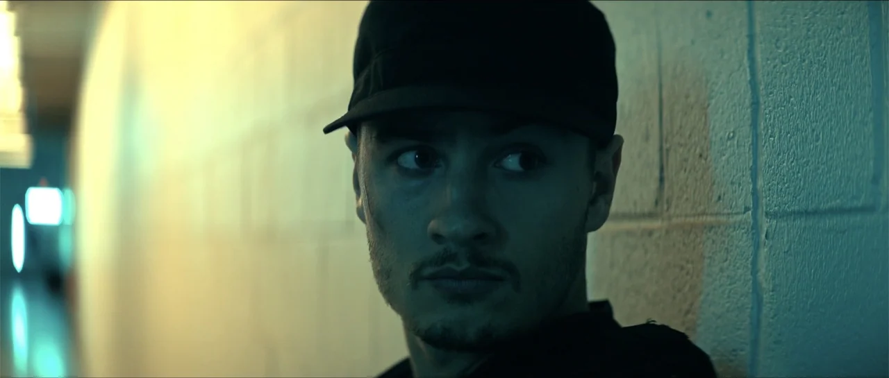 Cosmic Sin (2021) movie still: closeup — A dark hallway with a concrete brick wall features a man leaning back. The man wearing a cap looks sideways…