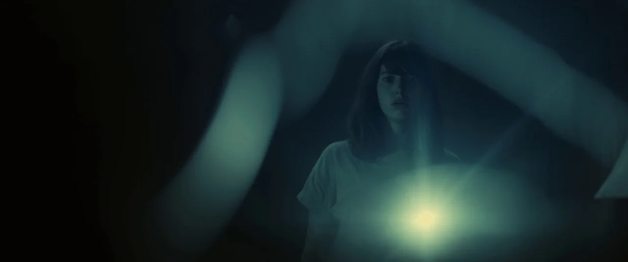 The Hole in the Ground (2019) movie still: medium — A woman stands in a dark environment with a single bright light illuminating her chest. Blurred…