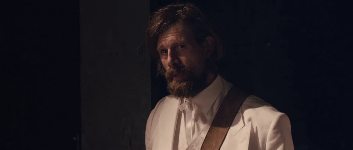 Broken Circle Breakdown (2012) movie still: medium — A man in a white suit stands within a dark, shadowed indoor space. The subject gazes towards…