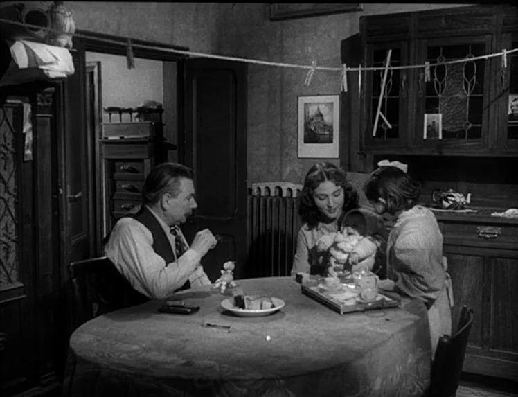 I Vitelloni (1953) movie still: medium — A domestic dining room with a wooden hutch and laundry line stretched above. A family sits around a covered…