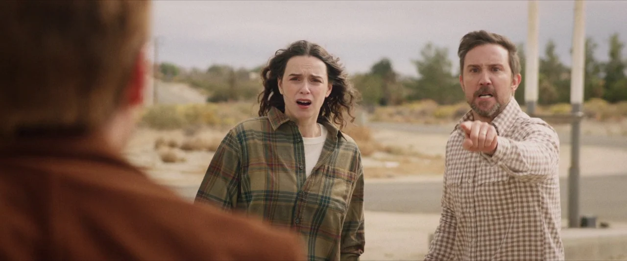 The Last Stop in Yuma County (2023) movie still: medium — A dry, desolate road sits under a flat overcast sky in the desert. A man in a gingham…