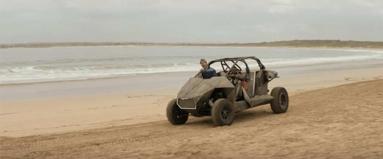 The Fall Guy (2024) movie still: wide — A wide, flat coastline features wet sand leading to a calm ocean under gray skies. Two men operate a custom…