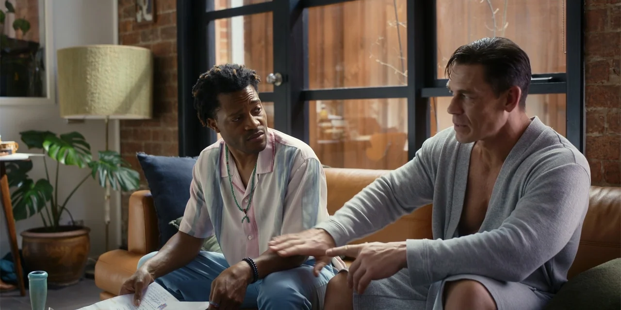 Ricky Stanicky (2024) movie still: medium — A modern living room features an exposed brick wall and a tan leather couch. Two men sit on the couch…