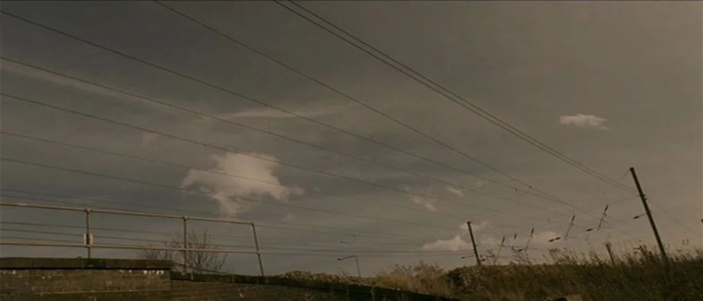 Another Year (2010) movie still: establishing — An outdoor railway track with overhead power lines against a dull, overcast sky. The scene shows a…