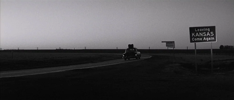 In Cold Blood (1967) movie still: wide — A flat rural landscape stretches under an evening sky at the state border. A sedan with luggage on its roof…