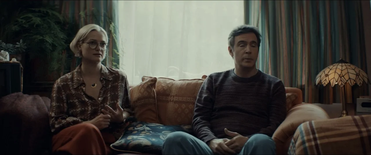 Bonus Track (2024) movie still: medium — A domestic space with patterned curtains and a floral sofa under a window. A woman and a man sit together…