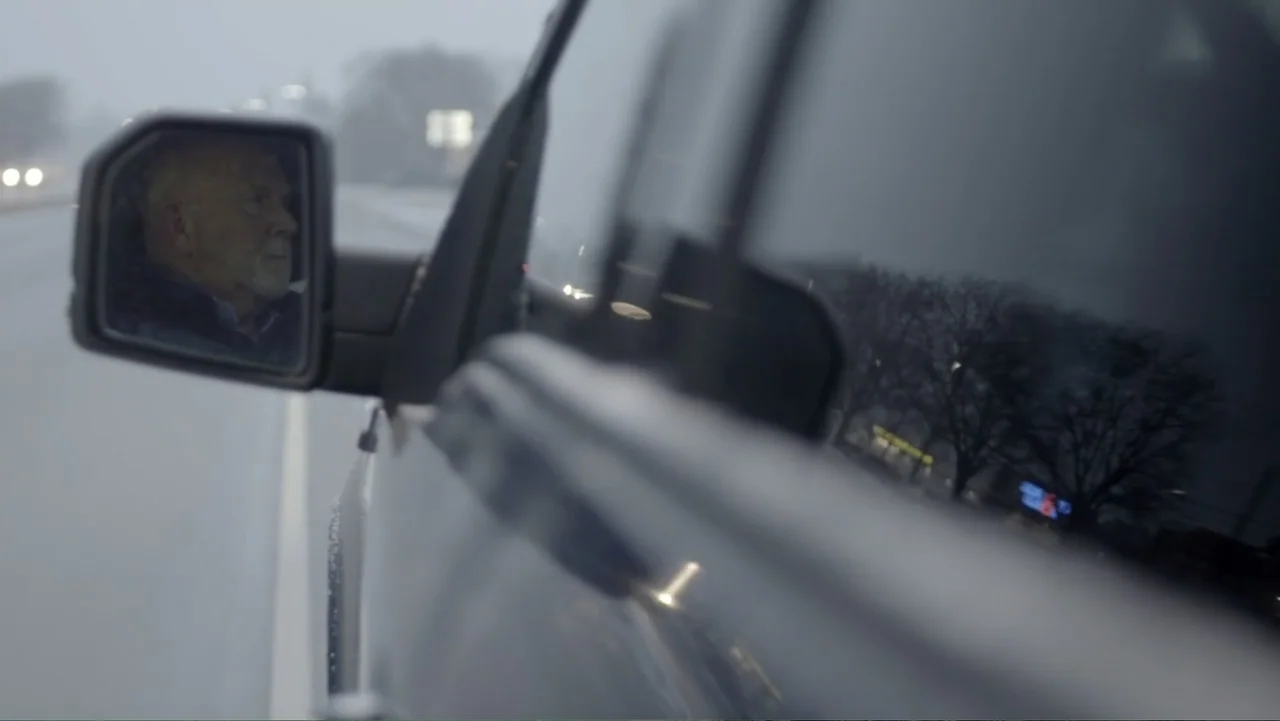 Murders at the Burger Joint (2022) movie still: closeup — A side mirror of a dark vehicle reflects an elderly man driving on a road. Bare trees and…