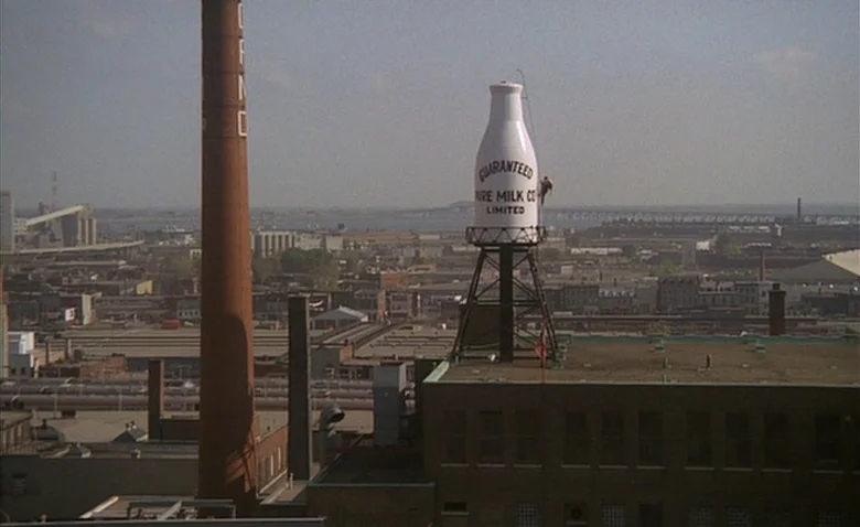 Sweet Movie (1974) movie still: wide — An industrial skyline features a massive white milk bottle water tower at its center. The foreground shows a…