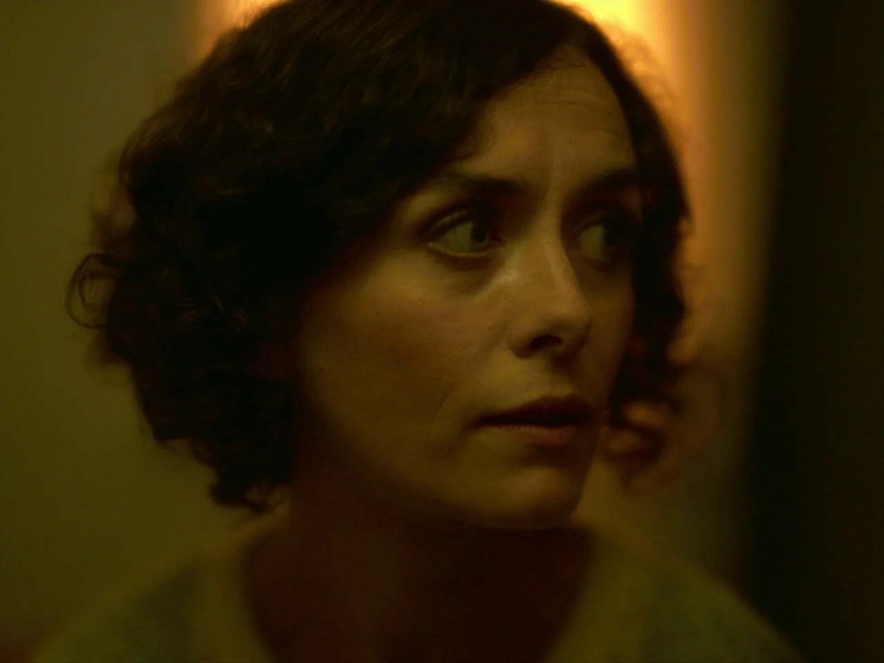 Violet (2014) movie still: closeup — A dimly lit interior space with a solid muted background. A woman with dark curly hair looks away from the…