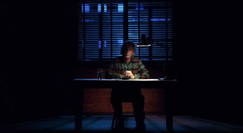 Gray’s Anatomy (1996) movie still: medium — An interrogation room with a dark wall and a window covered by metal blinds. A man sits at a bare table…