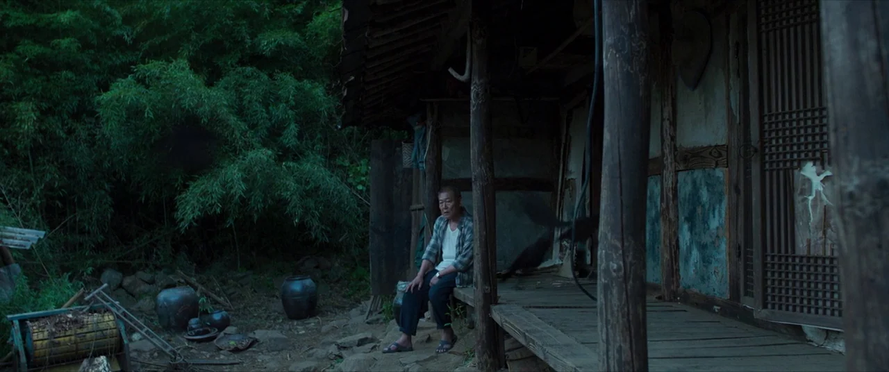 The Wailing (2016) movie still: wide — An elderly man sits on the wooden porch of a rustic, weathered rural home. Dense bamboo thickets surround the…