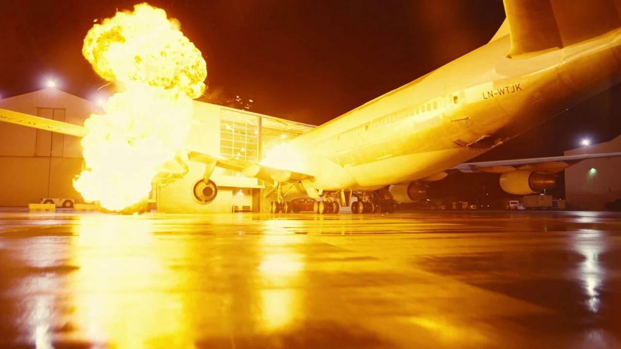 Tenet (2020) movie still: wide — An airport tarmac at night features a large passenger jet parked near terminal buildings. A massive, high-intensity…