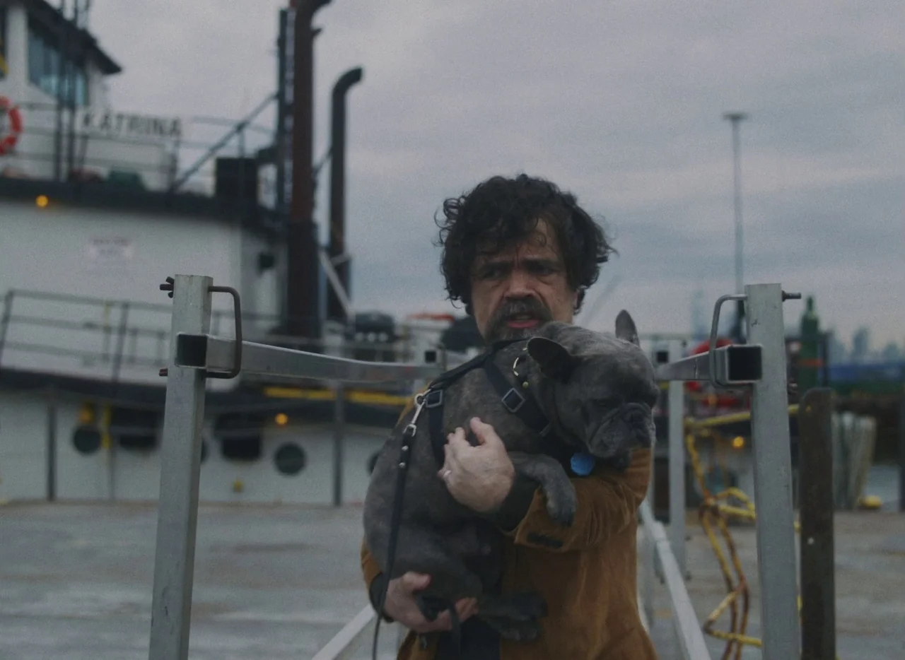 She Came To Me (2023) movie still: medium — A man stands on a grey concrete pier next to a moored tugboat. He is carrying a small grey French…