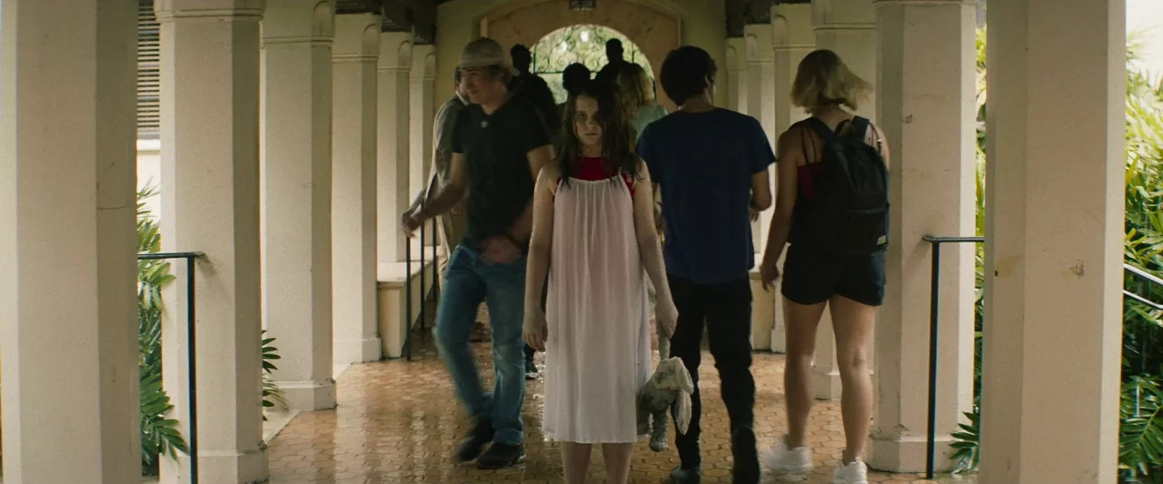 The Mad Hatter (2021) movie still: fullbody — A covered outdoor walkway with tall square columns leads to an arched doorway. A young girl stands…