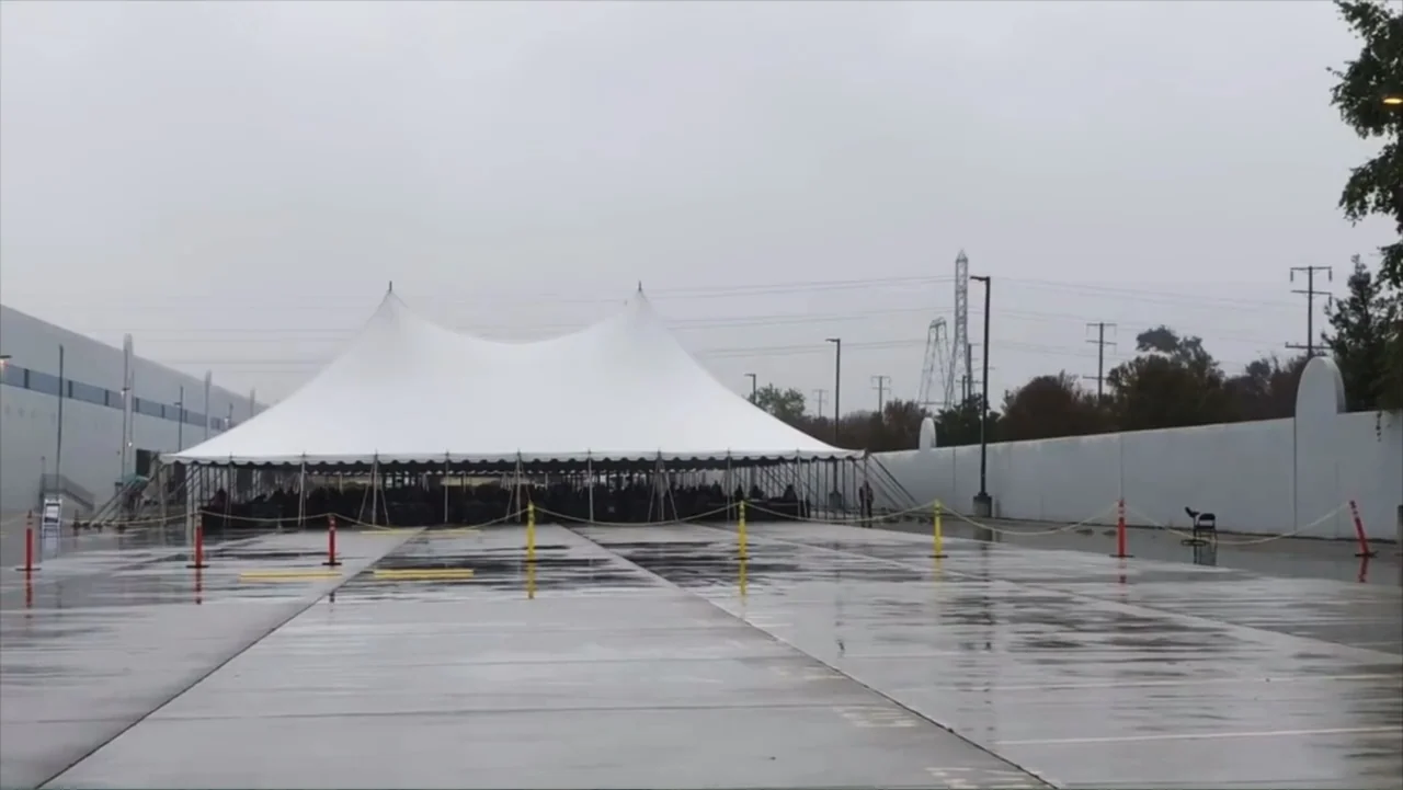 The Rise and Fall of Lularoe (2021) movie still: establishing — A wet asphalt parking lot under a cloudy gray sky features a large white tent. The…