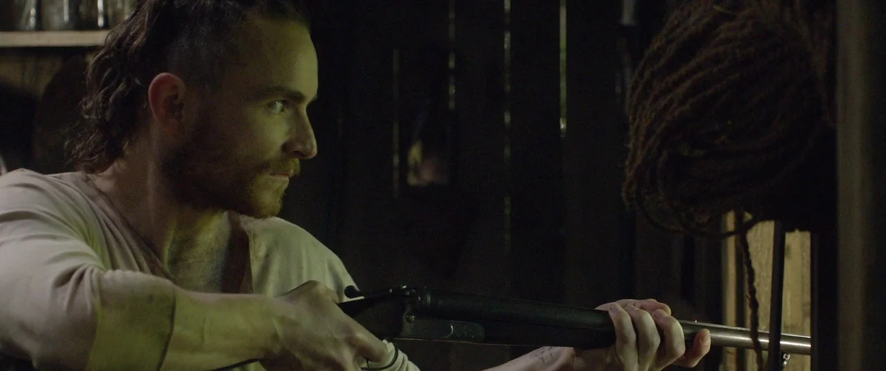 The Survivalist (2015) movie still: medium — A dimly lit interior space with dark wooden walls and textured wooden surfaces. A man grips a…