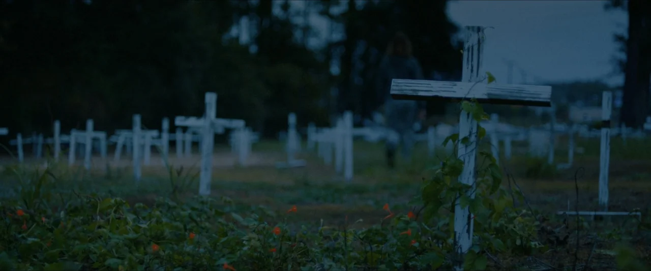 Ambush (2020) movie still: wide — A dark cemetery at dusk filled with rows of weathered white wooden crosses. A lone individual walks away from the…
