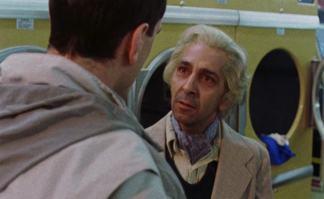 My Beautiful Laundrette (1985) movie still: medium — A 1970s laundromat features rows of yellow industrial drying machines. An older man speaks to a…