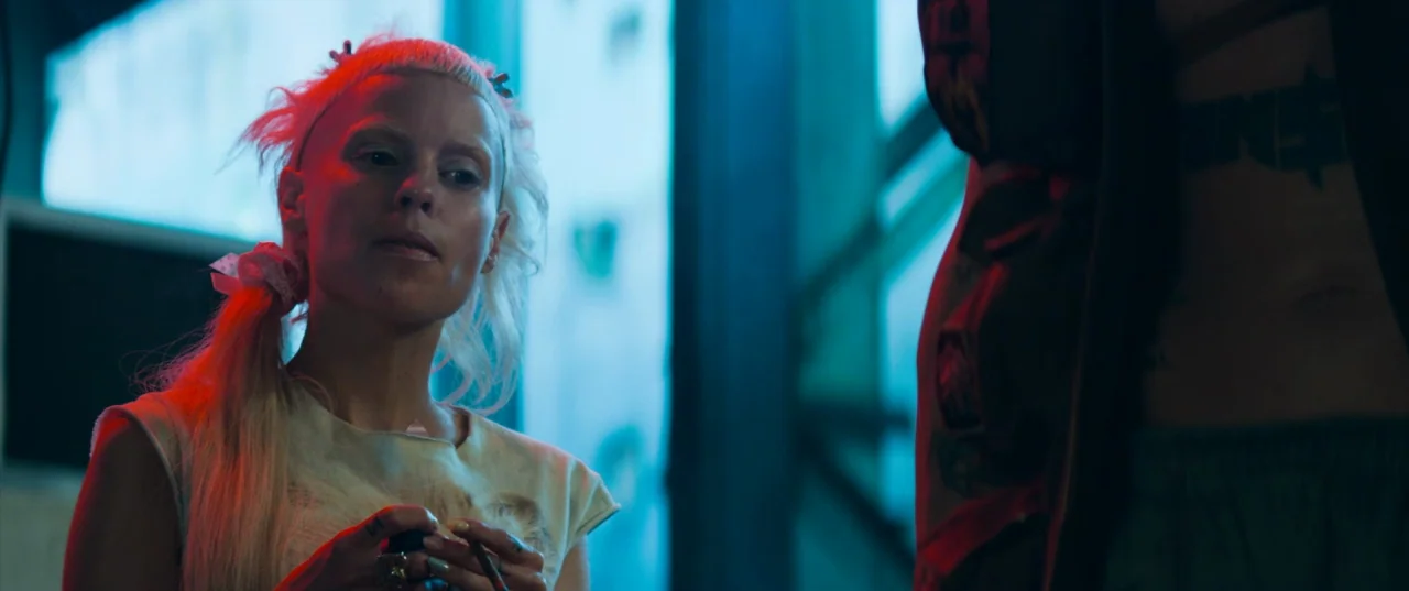 Chappie (2015) movie still: medium — An interior space features contrasting blue and red light sources hitting two people. A woman with bleached…