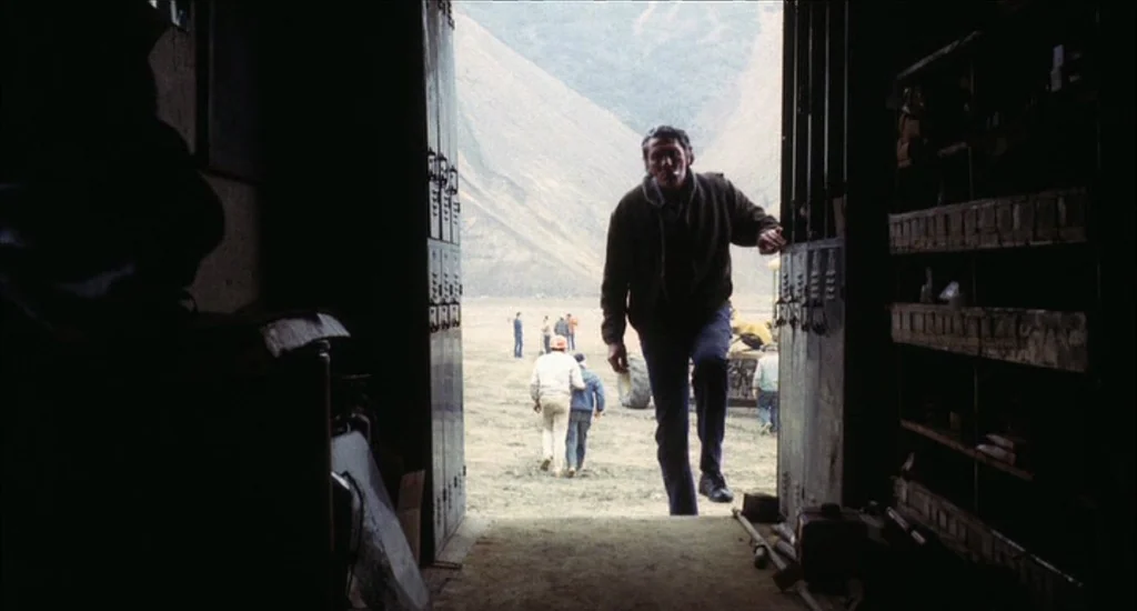 A Woman Under the Influence (1974) movie still: fullbody — A dark, industrial storage shed looks out onto a wide, grassy field with mountains. A man…