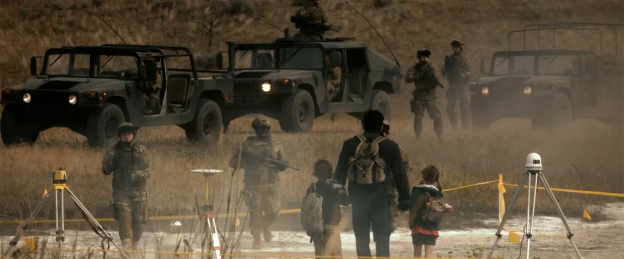 2012 (2009) movie still: wide — A dusty outdoor area contains camouflaged military vehicles and soldiers in tactical equipment. A man and two…