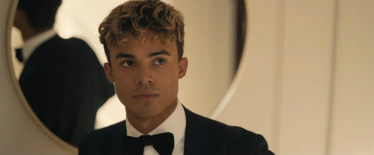My Fault: London (2025) movie still: closeup — A well-lit room contains a round mirror reflecting the back of a subject. A young man in a tuxedo…