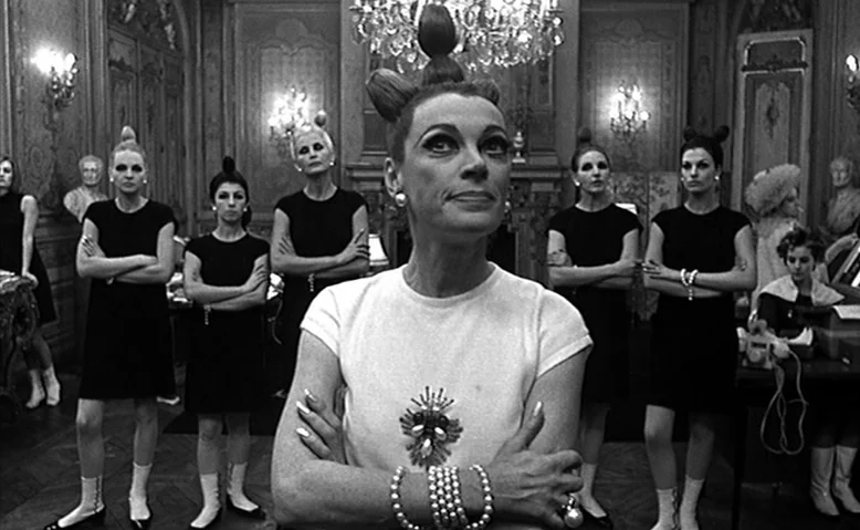 Who Are You, Polly Maggoo? (1966) movie still: medium — A group of women in 1960s fashion stand in an ornate, opulent room. The central subject, an…