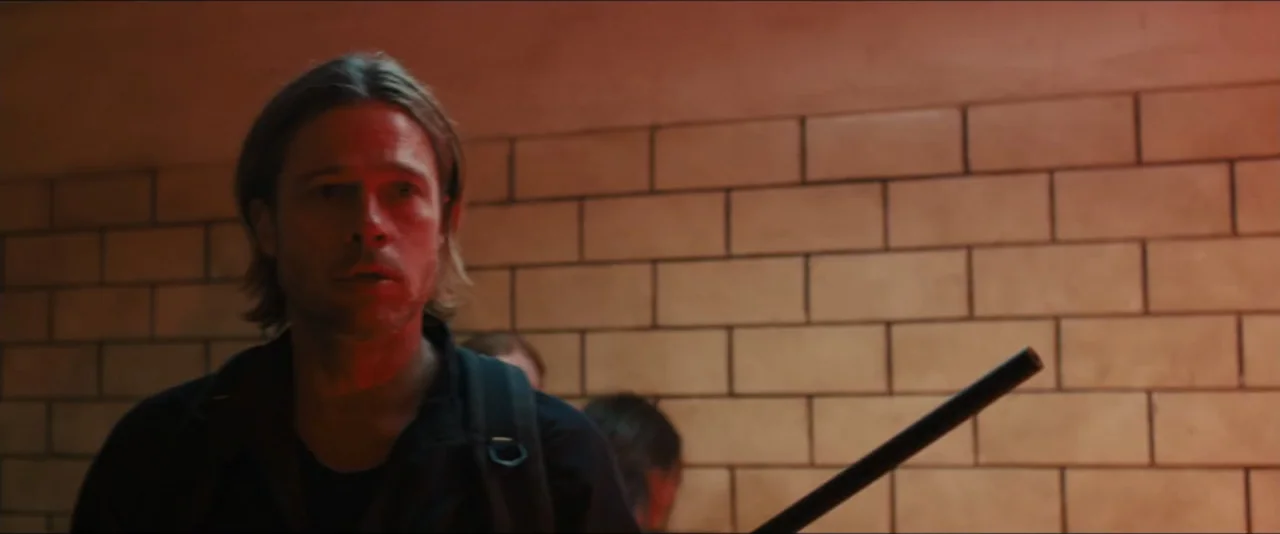 World War Z (2013) movie still: medium — A man stands in a dimly lit hallway with beige ceramic tiles. The subject holds a long thin dark object and…