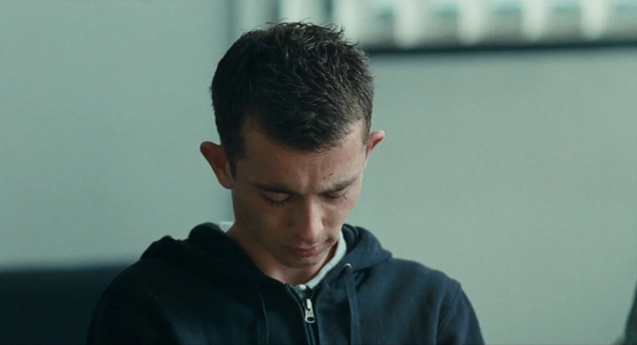 The Angel’s Share (2012) movie still: closeup — A plain interior with a pale grey wall provides a minimalist, institutional backdrop. A young man…