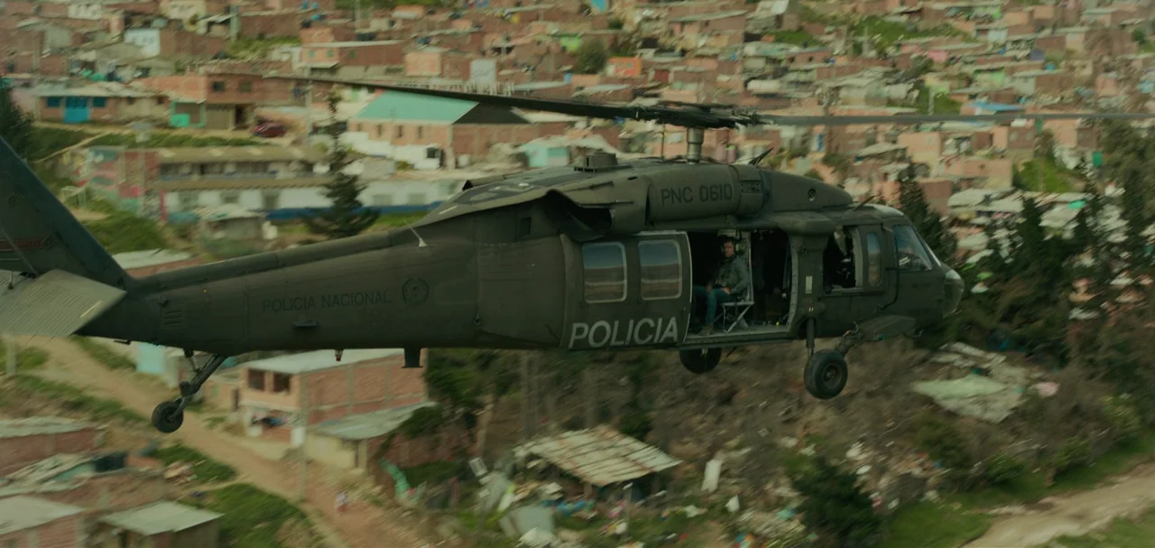 Triple Frontier (2019) movie still: wide — A military police helicopter flies over a densely populated hillside neighborhood of brick houses. A man…