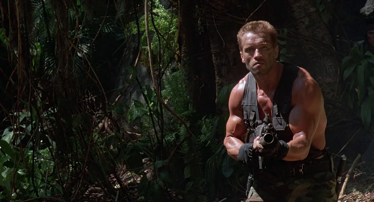 Predator (1987) movie still: medium — A dense, shadowed jungle environment filled with thick vines and dark foliage. A muscular man holds a tactical…