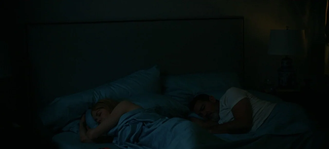 Babygirl (2024) movie still: medium — A darkened bedroom shows two people sleeping side by side under blue sheets. A lamp on the side table casts…