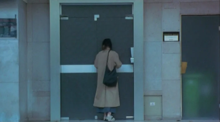Code Unknown (2000) movie still: fullbody — A person stands facing the closed double doors of a modern building facade. The structure features flat…