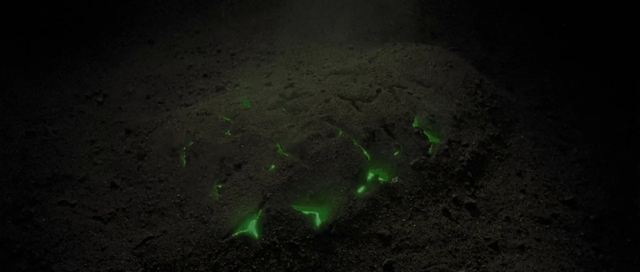Bones movie still: closeup — Dark, granular earth texture covers a patch of glowing material. Irregular fissures in the soil reveal a bright neon…